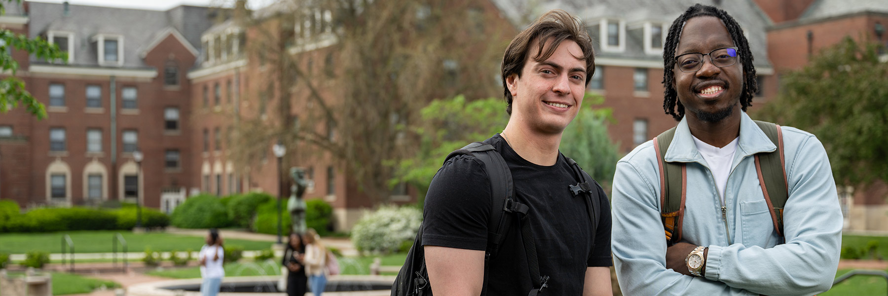 two male students with smiles pose on campus in front of building